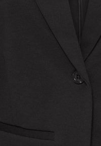 Zbliżenie na przód czarnego blezera z pojedynczym czarnym guzikiem zapiętym i wąską kieszenią wpuszczaną po lewej stronie.