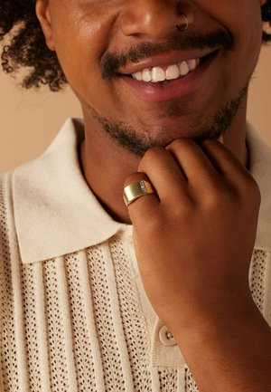 Gouden ring met een gladde, gebogen oppervlakte en een rij kleine, duidelijke accenten. Draagt op een hand die een crèmekleurig gebreid poloshirt aanraakt.
