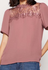 Růžový top s krátkými rukávy, s krajkovým panelem na výstřihu. Hladká textura, volný střih a jemné květinové krajkové detaily.
