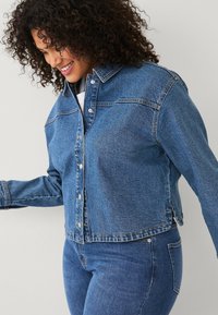 Giacca in denim di blu medio, con maniche lunghe, chiusura a bottoni e due tasche sul petto con dettagli di cucitura a contrasto.