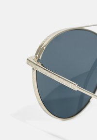 Lunettes de soleil rondes avec une monture dorée, des verres bleu foncé et des accents texturés sur les branches. Le design présente des détails gravés.
