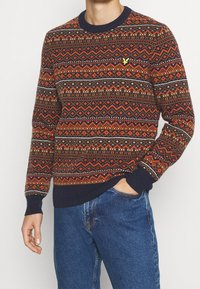 Man som bär en flerfärgad mönstrad tröja med marinblå muddar och krage, i kombination med blå jeans, med en hand i fickan, beskuren vy.