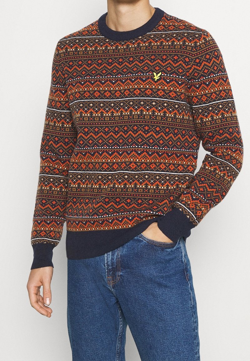 Man som bär en flerfärgad mönstrad tröja med marinblå muddar och krage, i kombination med blå jeans, med en hand i fickan, beskuren vy.