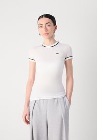 Weißes, kurzärmeliges, tailliertes T-Shirt mit grünen Ripp-Details am Kragen und an den Ärmeln, mit einem kleinen Lacoste-Logo auf der linken Brust.