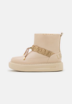 Botte beige mi-mollet avec semelle en caoutchouc épaisse, sangle froncée avec accents dorés à l'avant, et languette de tirage à l'arrière.