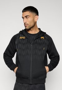 Μαύρο φούτερ με φερμουάρ και γεωμετρικό υφασμάτινο σχέδιο, με κίτρινες λεπτομέρειες και λογότυπο. Σχεδιασμένο με κουκούλα και τσέπες, κατασκευασμένο από απαλό υλικό.