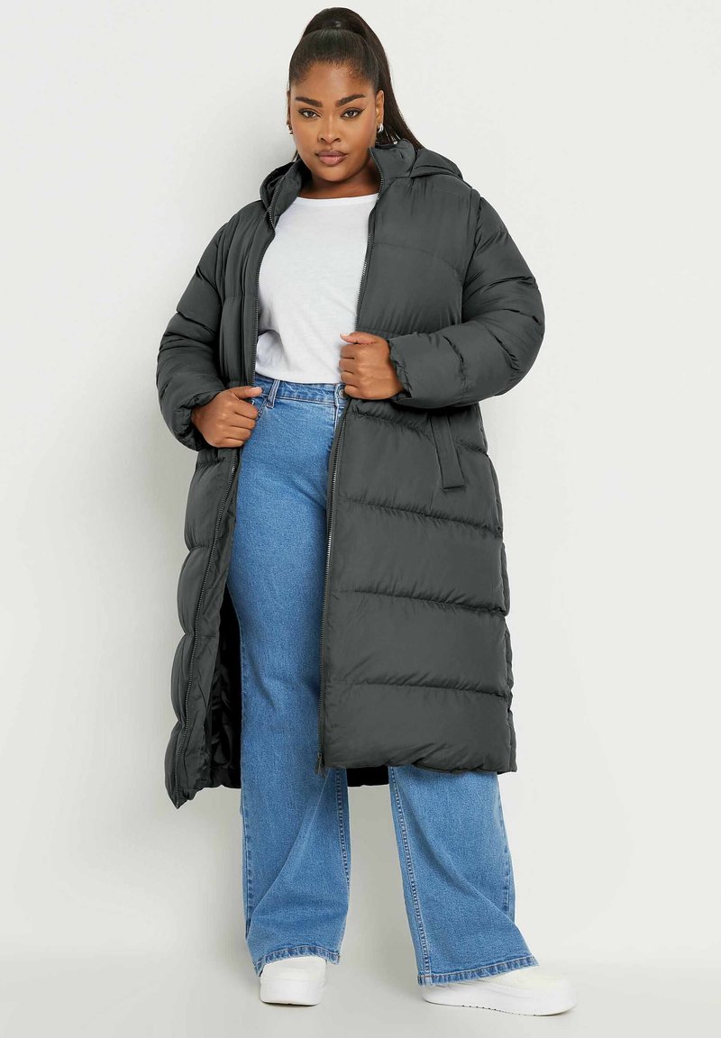 Abrigo tipo puffer en gris oscuro con capucha, de largo largo, diseño con acolchado vertical y cremallera frontal. Combinado con jeans azul claro y zapatos blancos.
