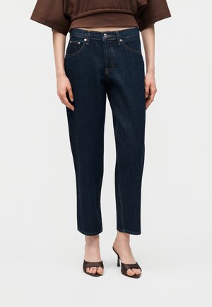 Femme portant un jean droit bleu foncé et des sandales à talons brun ouvertes à l'avant, debout devant un fond clair uni.