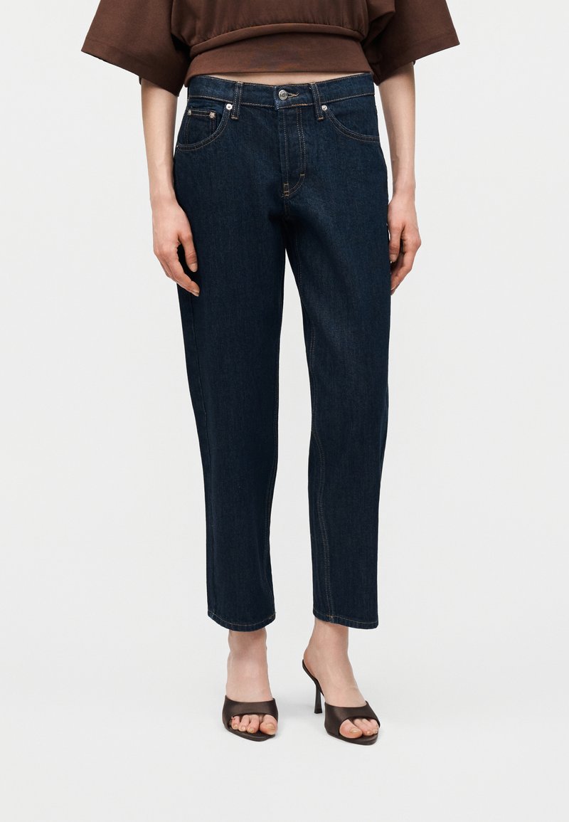 Γυναίκα που φοράει σκούρα μπλε τζιν με ίσια μπατζάκια και καφέ σανδάλια με τακούνι και ανοιχτά δάχτυλα, στέκεται μπροστά σε απλό ανοιχτό φόντο.