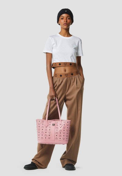 Giovane donna che indossa un crop top bianco, pantaloni marroni a gamba larga, berretto nero, tiene una borsa tote rosa decorata, in piedi davanti a uno sfondo neutro.