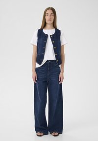 Farmerdzseki öt nagy gombbal, fehér pólóval és bő szárú farmernadrággal párosítva. Sötétkék, sima textúrájú anyag.