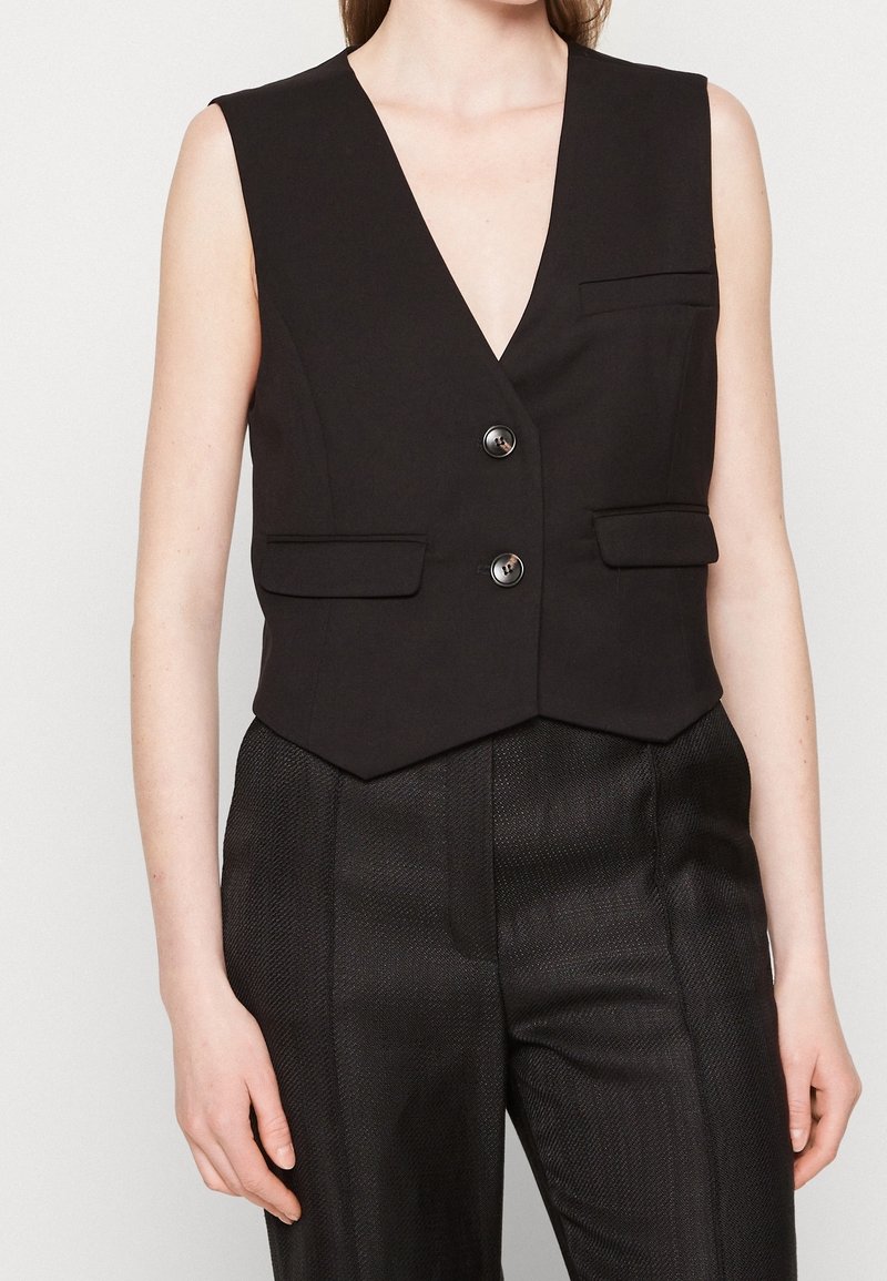 Donna che indossa un gilet nero senza maniche con tasche a patta e pantaloni neri su misura con texture, su uno sfondo neutro.