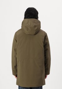 Parka imperméable vert olive avec une grande capuche, des manches longues et une coupe droite. Texture lisse avec un design minimaliste et sans matériel visible.
