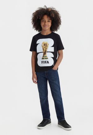 Enfant aux cheveux bouclés portant un t-shirt noir FIFA Coupe du Monde 2026, un jean bleu et des baskets noires, debout avec une main dans la poche.