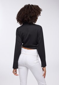 Blusa corta in satin nero con maniche lunghe, abbinata a jeans bianchi aderenti con un dettaglio decorativo a catena in vita.