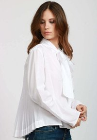 Blusa bianca con colletto a fiocco, design con pieghe sulla parte posteriore, maniche lunghe e texture liscia. Indossata con jeans blu.