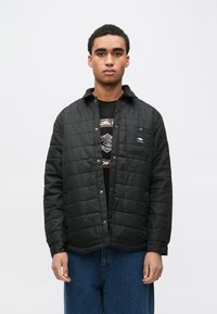 Czarna pikowana kurtka z zapięciem na guziki z przodu, kieszenią na piersi i kołnierzem, noszona na koszulce z nadrukiem. Charakteryzuje się luźnym krojem i fakturowaną tkaniną.