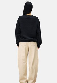 Vrouw met lang zwart haar, gekleed in een losse zwarte sweatshirt en beige wijde broek, staand met haar rug naar de camera tegen een effen achtergrond.