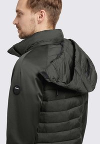 Dunkelgrüne Jacke mit einem geformten Kragen, gestepptem Körper und gepolstertem Kapuze mit verstellbarem Kordelzug. Enthält ein Logopatch an der Schulter.