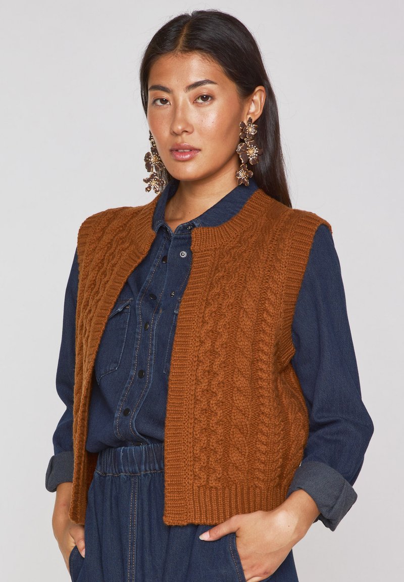 Gilet en tricot marron avec un motif de câbles texturé, arborant un col rond, porté par-dessus une chemise en denim foncé. De grandes boucles d'oreilles florales complètent le look.
