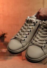 Weiße Ledersneaker mit mehrfarbigen Ösen, die von einer Hand vor einem unscharfen, warmtonigen Hintergrund mit einer kleinen Topfpflanze gehalten werden.