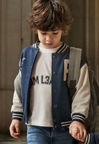 Giacca varsity blu navy con maniche grigie, caratterizzata da un colletto a righe bianche e dalla lettera "P" in evidenza. Indossata sopra una maglietta bianca con scritte.