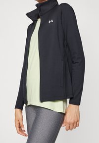 Svart zip-up sportjacka med hög krage, tillverkad av lätt material. Har logotyp på bröstet och sidofickor. Lagd över en ljusgrön topp.