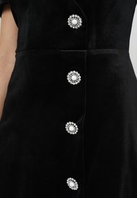 Czarna aksamitna sukienka z pięcioma okrągłymi, ozdobionymi guzikami z białymi środkami i srebrnymi akcentami, tworząca teksturowany i elegancki wygląd.
