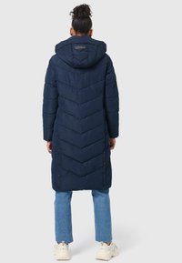 Longue doudoune bleu marine avec un motif en chevrons, une capuche et des accents en boutons. Associée à un jean bleu clair et des baskets blanches.