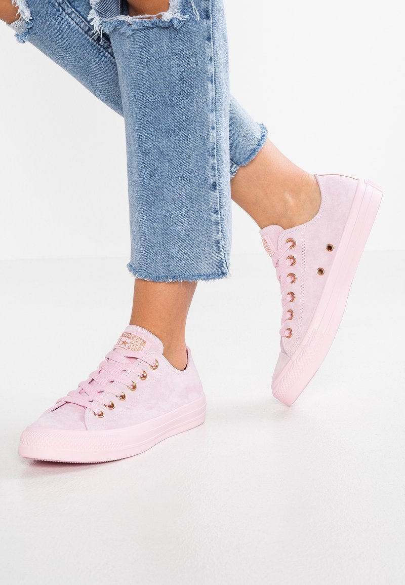 Sneakers in suede rosa chiaro con punta rotonda, suola piatta e occhielli dorati. Abbinati a jeans blu strappati, che mostrano orli sfilacciati.