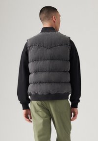 Sivi puffer prsluk s rebrastim ovratnikom, šavnim detaljima i opuštenim krojem, nosi se preko crne dukserice i zelenih hlača.