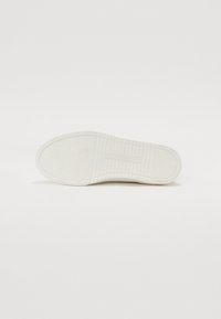 Hvite gummisåler til sneakers med en teksturert overflate, som har et sekskantet mønster og horisontale spor for bedre grep. Merkevaren er synlig.