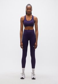 Γυναίκα όρθια, φορώντας σκούρο μοβ αθλητικό τοπ και κολάν, λευκές κάλτσες και λευκά αθλητικά παπούτσια, μπροστά σε λευκό φόντο.