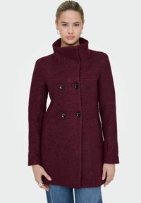 Burgundijas vilnas mēteli ar augstu apkakli, dubultā pogu dizainu un melnām pogām. Iezīmējas ar teksturētu audumu un gariem piedurknēm.