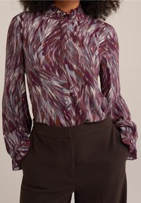 Long-sleeved blouse with a ruffled collar, featuring a violet and cream abstract pattern. Fabric is lightweight with slight sheen.