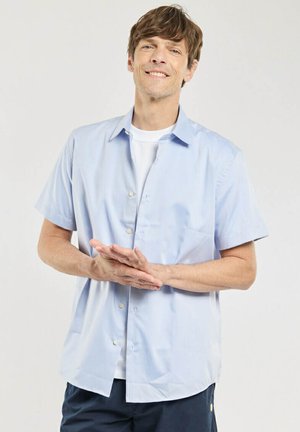 Homme souriant aux cheveux châtain clair portant une chemise à manches courtes bleu clair boutonnée sur un t-shirt blanc, mains jointes devant, fond neutre.