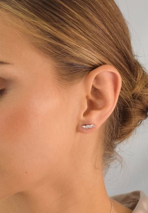 Silberne blattförmige Ohrringe mit aufwendigen Details und kleinen Edelsteinen, vor einem hellen Hintergrund getragen, nah am Ohr.