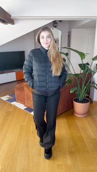 Juoda pūkinė striukė su aukštu apykakle, užtrauktuko uždarymu ir ilgomis rankovėmis, derinama su juodomis kelnėmis ir juodais batais, interjere.