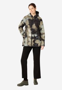 Chaqueta acolchada con capucha y un patrón de hojas multicolores en tonos verdes, negros y beiges. Combinada con pantalones negros y zapatos verdes.
