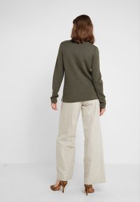 Pull à manches longues vert olive avec un col rond, associé à un pantalon beige ample et des talons hauts rayés. Tissu en maille texturée.