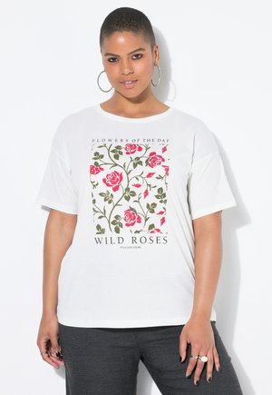 Femme aux cheveux courts portant des boucles d'oreilles créoles et un t-shirt blanc avec un motif de rose sauvage rose et verte, ainsi que des pantalons noirs, se tenant devant un fond blanc.