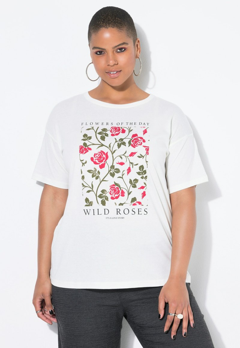 Femme aux cheveux courts portant des boucles d'oreilles créoles et un t-shirt blanc avec un motif de rose sauvage rose et verte, ainsi que des pantalons noirs, se tenant devant un fond blanc.