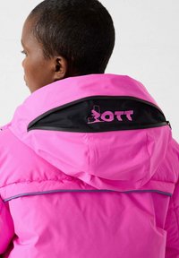 Persona che indossa una giacca rosa con cappuccio caratterizzato da un pannello nero e un logo da sci rosa con testo sul retro.