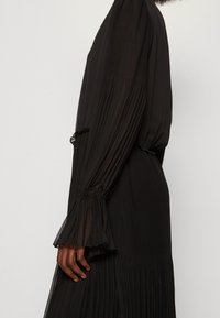 Svart långärmad klänning med veckad design, åtsittande midja och utsvängda muddar. Lättvikts- och texturerat tyg med en flödande silhuett.