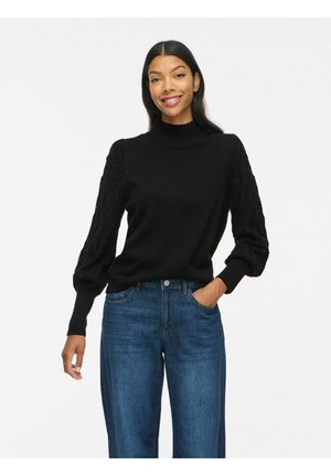 Femme aux longs cheveux foncés portant un pull noir avec des manches bouffantes texturées et un jean bleu, souriant avec une main dans la poche.