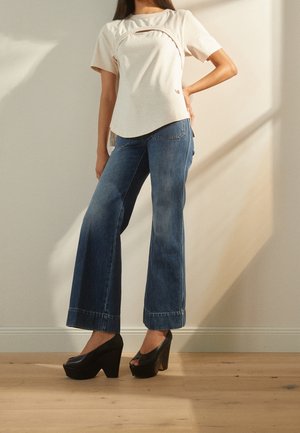 Kvinde iført højtaljede blå vidde-ben jeans, beige kortærmet top og sorte platform-højhælede sko står indendørs.