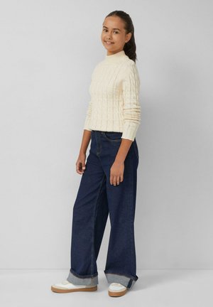 Cremefarbener Grobstrickpullover mit hohem Kragen, kombiniert mit locker sitzenden, dunklen Denim-Jeans mit hochgerollten Säumen. Weiße Sneakers mit Gummisohlen.