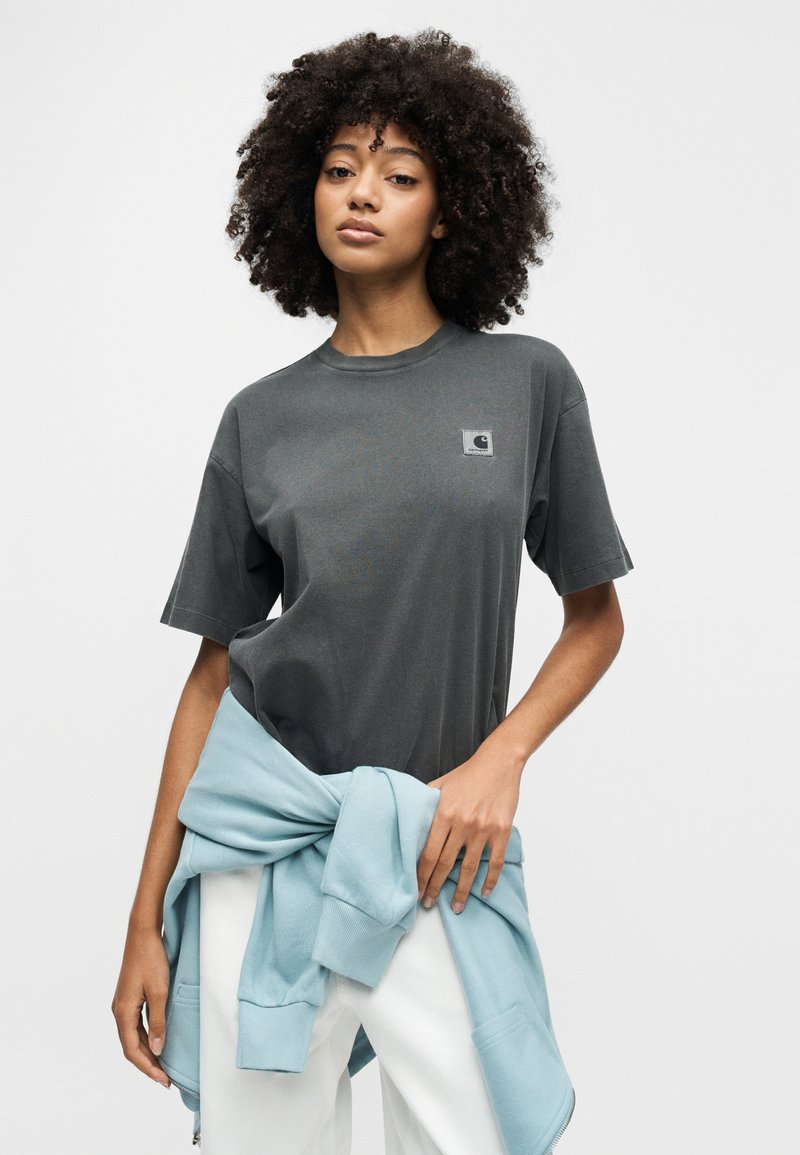 Giovane donna con capelli ricci che indossa una t-shirt Carhartt grigio scuro e una felpa azzurra legata in vita.