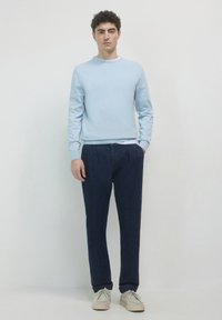 Helltürkises Sweatshirt mit Rundhalsausschnitt, dunkelblaue plissee Hosen und beigefarbene Schnürschuhe. Weiches Material, taillierte Passform und entspannte Silhouette.