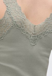 Γκρι ριμπ αμάνικη μπλούζα με δαντέλα με λουλούδια κατά μήκος της λαιμόκοψης και του στήθους σε φόντο γυμνής επιδερμίδας.